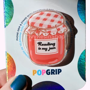 May include: A PopGrip phone grip with a red and white gingham-patterned jar design and the text "Reading is my jam." The phone accessory is designed to grip and stand with a phone.