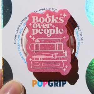 Puede incluir: Agarre para teléfono rosa con un acabado brillante y un diseño blanco que dice "Books over people" con una pila de libros debajo del texto.