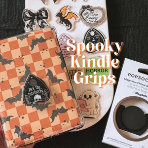 May include: A Kindle case with an orange and cream checkered pattern featuring black bats and a black planchette-shaped grip labeled Hex the Patriarchy. Nearby are various acrylic charms and a black magnetic PopSockets phone grip and stand.
