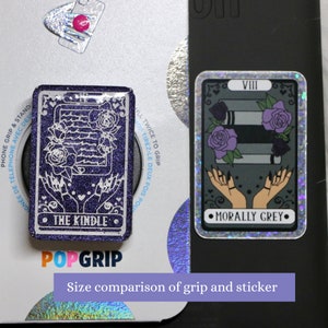 May include: A purple glitter phone grip with the text "THE KINDLE" and a white outline of a phone and hands. The grip is next to a sticker with a gray background and the text "MORALLY GREY" and a white outline of hands and a stack of books. The sticker has a holographic finish. The text "VIII" is at the top of the sticker. The text "Size comparison of grip and sticker" is at the bottom of the image.
