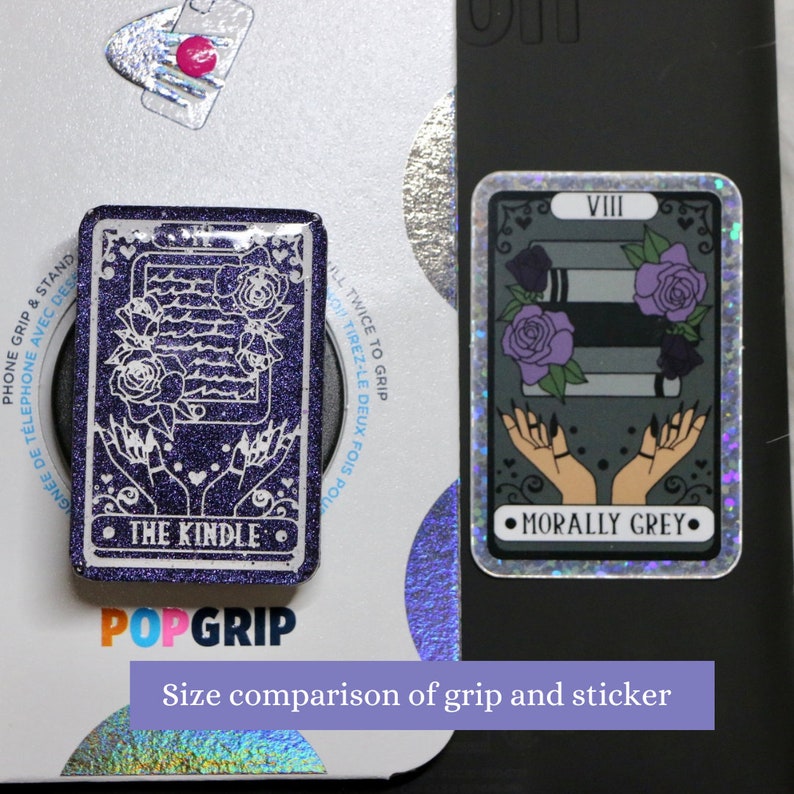 May include: A purple glitter phone grip with the text "THE KINDLE" and a white outline of a phone and hands. The grip is next to a sticker with a gray background and the text "MORALLY GREY" and a white outline of hands and a stack of books. The sticker has a holographic finish. The text "VIII" is at the top of the sticker. The text "Size comparison of grip and sticker" is at the bottom of the image.