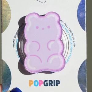 May include: A light purple PopGrip phone grip in the shape of a gummy bear. The phone grip has a glittery border and is attached to a white and iridescent background. The words "PHONE GRIP & STAND" and "POPGRIP" are visible.