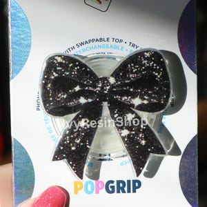 May include: Black bow-shaped PopGrip with glitter and star accents. The PopGrip has a swappable top and is attached to a white base. The text "POPGRIP" is visible at the bottom.