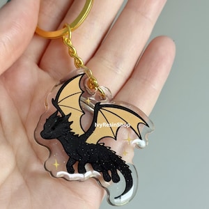 May include: A black dragon keychain with gold wings and a gold chain. The dragon has a detailed design and is set against a clear background with gold stars. The keychain is from IvyResinShop.