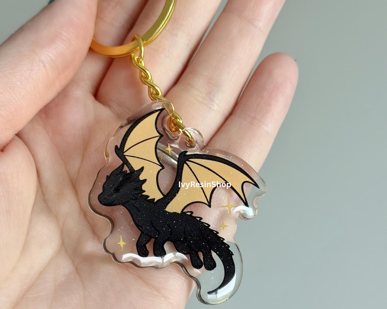 May include: A black dragon keychain with gold wings and a gold chain. The dragon has a detailed design and is set against a clear background with gold stars. The keychain is from IvyResinShop.