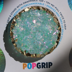 May include: A turquoise blue and gold PopGrip phone grip with a glittery finish. The text "PopGrip" is printed in pink, blue, and black letters on the white background.