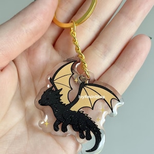 May include: A black dragon keychain with gold wings and a gold-colored keyring. The dragon is outlined in black and has a glittery texture. The keychain is held in a hand.