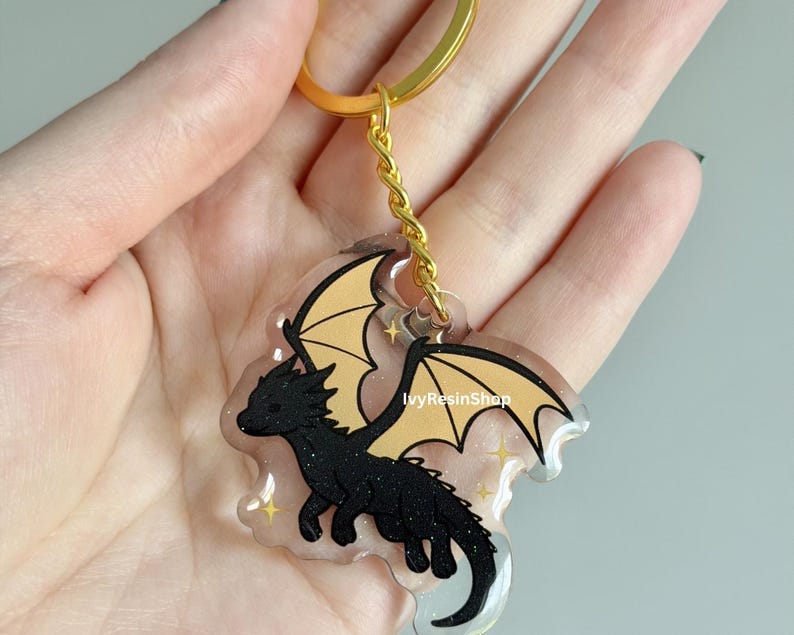 May include: A black dragon keychain with gold wings and a gold-colored keyring. The dragon is outlined in black and has a glittery texture. The keychain is held in a hand.