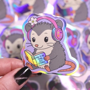 May include: Holographic sticker featuring a cartoon opossum wearing headphones and reading a book. The book's cover reads "How to be Feral but Literate." The opossum has a flower on its head and a purple background.
