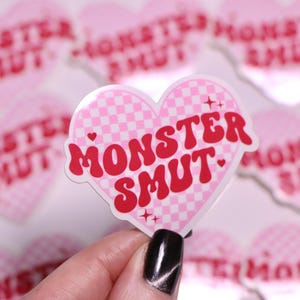 K&ouml;nnte beinhalten: Ein rosa herzf&ouml;rmiger Aufkleber mit einem Karomuster und den Worten "MONSTER SMUT" in Rot. Der Aufkleber hat einen wei&szlig;en Rand und kleine Sternakzente. Weitere &auml;hnliche Aufkleber befinden sich im Hintergrund.