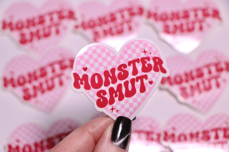 K&ouml;nnte beinhalten: Ein rosa herzf&ouml;rmiger Aufkleber mit einem Karomuster und den Worten "MONSTER SMUT" in Rot. Der Aufkleber hat einen wei&szlig;en Rand und kleine Sternakzente. Weitere &auml;hnliche Aufkleber befinden sich im Hintergrund.