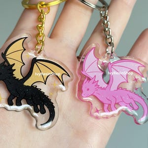May include: Two dragon-shaped keychains. One is black with yellow wings, the other is pink with pink wings. Both have a clear border with glitter and the text "IvyResinShop". Each keychain has a metal ring and chain.