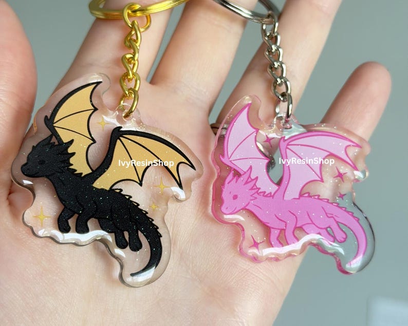 May include: Two dragon-shaped keychains. One is black with yellow wings, the other is pink with pink wings. Both have a clear border with glitter and the text "IvyResinShop". Each keychain has a metal ring and chain.