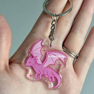 May include: A pink dragon keychain with a silver keyring and chain. The dragon has outstretched wings and is outlined in a darker pink. The keychain has a clear background with glitter and star accents. The text "IvyResinShop" is visible.