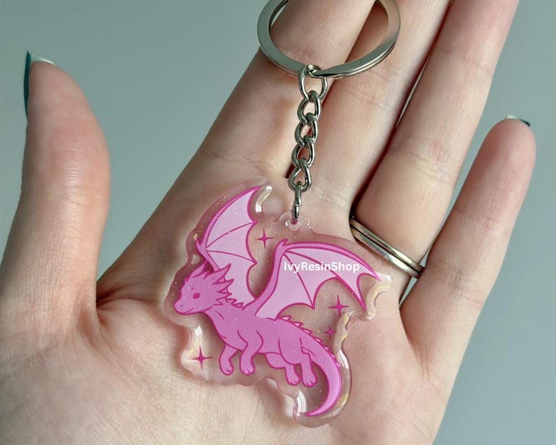 May include: A pink dragon keychain with a silver keyring and chain. The dragon has outstretched wings and is outlined in a darker pink. The keychain has a clear background with glitter and star accents. The text "IvyResinShop" is visible.