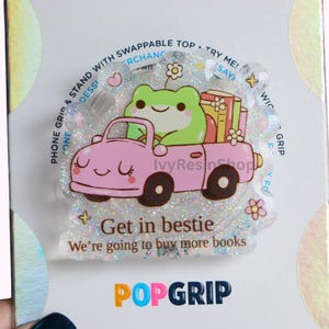Könnte beinhalten: Ein PopGrip-Telefonhalter mit einem rosa Auto, das von einem lächelnden Frosch gefahren wird und Bücher transportiert. Der Text "Get in bestie, we're going to buy more books" steht darunter. Der Halter hat einen klaren, glitzernden Hintergrund.