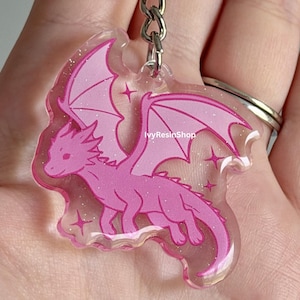 May include: A pink dragon keychain with glitter accents. The dragon has large wings and is surrounded by star shapes. The keychain is attached to a silver metal ring.