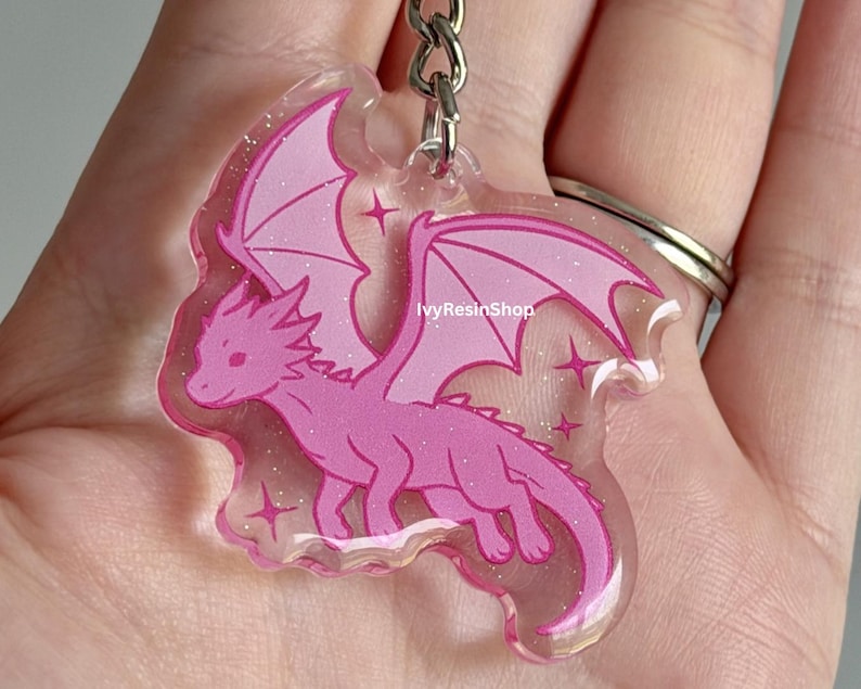 May include: A pink dragon keychain with glitter accents. The dragon has large wings and is surrounded by star shapes. The keychain is attached to a silver metal ring.