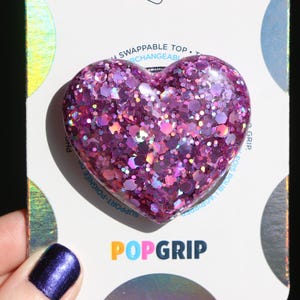 May include: A heart-shaped PopGrip with a glittery purple design. The PopGrip is on a white card with the PopGrip logo in orange and the words "SWAPPABLE TOP" printed on it.