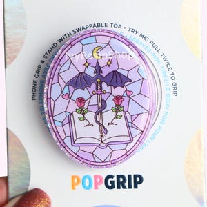 以下が含まれることがあります： 紫とピンクのPopGripフォンスタンド。ステンドグラスのデザインで、ドラゴン、剣、開いた本、バラ、ハート、三日月が描かれています。PopGripはキラキラした縁取りで、「POPGRIP」の文字が入っています。