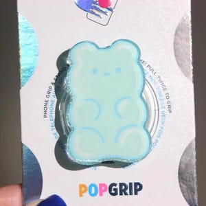 May include: A light blue gummy bear PopGrip phone stand. The phone grip has a glittery border and is on a white background with the words "PHONE GRIP & STAND" and "POPGRIP".