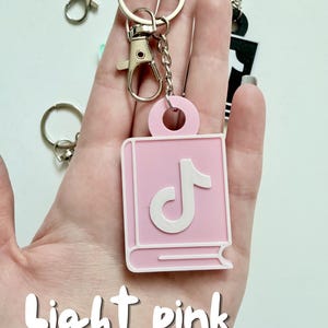 May include: Light pink keychain featuring a book-shaped design with a white musical note symbol. The keychain is attached to a silver metal clasp and ring. The words "Light pink" are written in a script font at the bottom of the image.