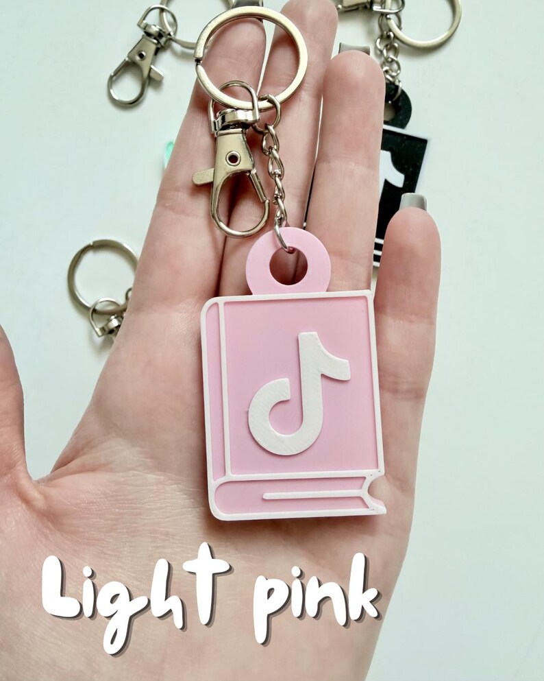 May include: Light pink keychain featuring a book-shaped design with a white musical note symbol. The keychain is attached to a silver metal clasp and ring. The words "Light pink" are written in a script font at the bottom of the image.