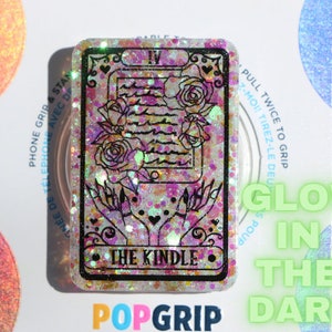 May include: A pink and white glitter phone grip with a black outline of a tarot card featuring a rose and the text "The Kindle". The grip glows in the dark.