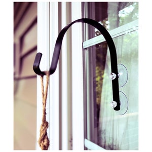 May include: Black metal hook with a suction cup attachment for hanging items on a window. The hook is attached to a window with two suction cups.