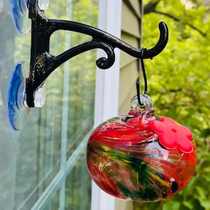 May include: A decorative glass hummingbird feeder with a vibrant red, green, and yellow design. The feeder is suspended from a black metal bracket attached to a window with suction cups. A red flower-shaped perch is attached.