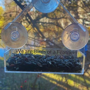 May include: A clear plastic bird feeder with suction cups attached to a window. The feeder has a small tray filled with birdseed and the text "We are Birds of a Feather" printed on the front.