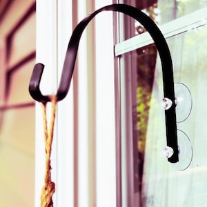 May include: A black metal hook with a suction cup attachment for hanging items on a window. The hook is attached to a window with two suction cups. A rope is hanging from the hook.
