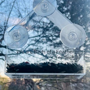 May include: A clear plastic bird feeder with suction cups, attached to a window. The feeder has a small tray filled with birdseed and the text "We are birds of a feather Love Sarah" is printed on the front.