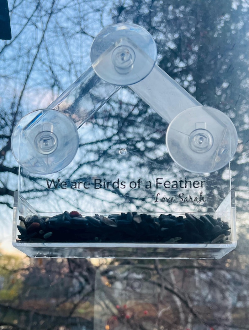 May include: A clear plastic bird feeder with suction cups, attached to a window. The feeder has a small tray filled with birdseed and the text "We are birds of a feather Love Sarah" is printed on the front.