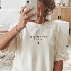 May include: An off-white short-sleeve t-shirt featuring the black text "GROWING A TINY human" centered on the chest above a small black heart icon. The shirt has a crew neckline and a relaxed fit, made of a solid-colored fabric.