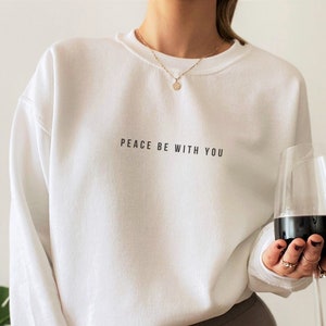 Pu&ograve; includere: Felpa bianca con la scritta "PEACE BE WITH YOU" stampata sul davanti. La modella indossa leggings marroni con un logo bianco.