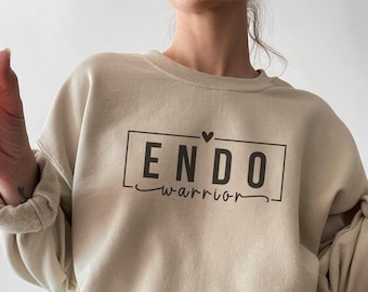 May include: A beige sweatshirt with the text "ENDO warrior" in black lettering inside a black rectangle. A small black heart is above the text.
