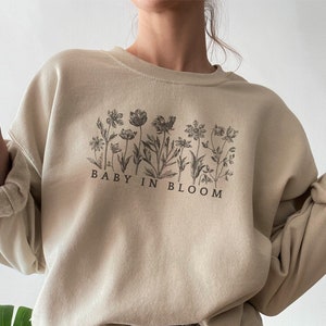 May include: A beige sweatshirt with a floral graphic and the text "BABY IN BLOOM" printed on the front.