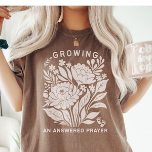 May include: An espresso-colored short-sleeve t-shirt featuring a large white graphic of a floral bouquet. The text "GROWING" appears above the flowers, and "AN ANSWERED PRAYER" is printed below, accompanied by two small baby footprint icons.