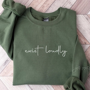 May include: A green sweatshirt with the text "exist loudly" printed in white on the front. The sweatshirt is folded and laid on a wooden surface.