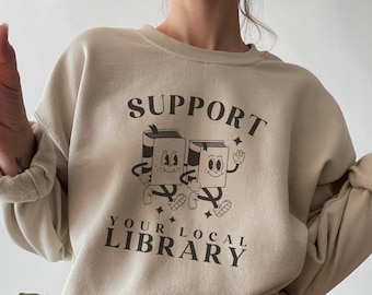 May include: A beige sweatshirt with the text "SUPPORT YOUR LOCAL LIBRARY" and a graphic of two cartoon books with faces.