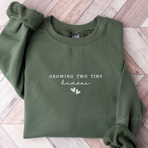May include: Military green sweatshirt with the text "GROWING TWO TINY humans" and two small hearts. The long-sleeved top is made of a soft material and is laid flat on a wooden surface.
