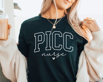 May include: A black t-shirt with white text that reads "PICC nurse" with a small heart symbol. The shirt is being worn by a person sitting on a couch.