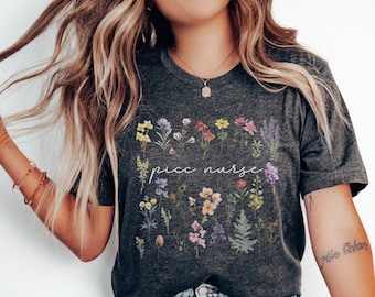 May include: A dark grey t-shirt with a floral design and the text "PICC nurse" in white script. The shirt is being worn by a person with long blonde hair and blue jeans.