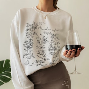May include: White sweatshirt with a floral design and the word "Midwife" printed on the front. The sweatshirt is being worn by a person with a glass of red wine in their hand.