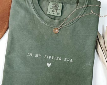 May include: A green t-shirt with the text "IN MY FIFTIES ERA" and a small heart graphic. The shirt is made of a soft, comfortable material.