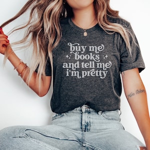 May include: A dark heather gray short-sleeve t-shirt featuring the white text "buy me books and tell me i'm pretty" with small star accents. The shirt has a crew neckline and is paired with light blue denim jeans that have a distressed knee detail.