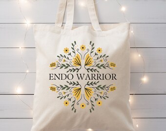 May include: A cream-colored canvas tote bag featuring two long shoulder straps. The center of the bag displays the text ENDO WARRIOR in black capital letters, surrounded by a symmetrical floral design with yellow flowers and green leaves.