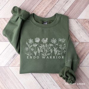 Pu&ograve; includere: Una felpa verde con un disegno floreale bianco e il testo "ENDO WARRIOR".