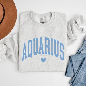 Puede incluir: Una sudadera gris con un gr&aacute;fico azul "AQUARIUS" en una fuente arqueada con un peque&ntilde;o coraz&oacute;n azul debajo.